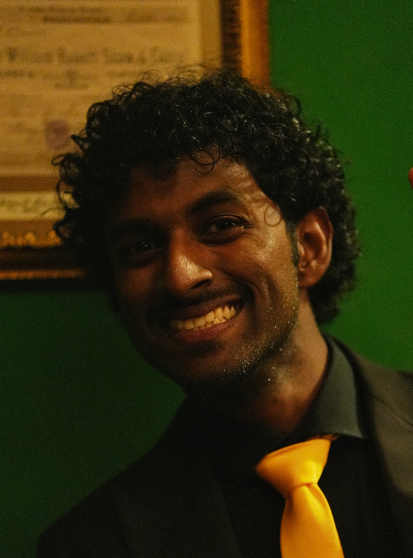 Portrait of Ravike Leon, smiling, in a dark suit with a gold tie
